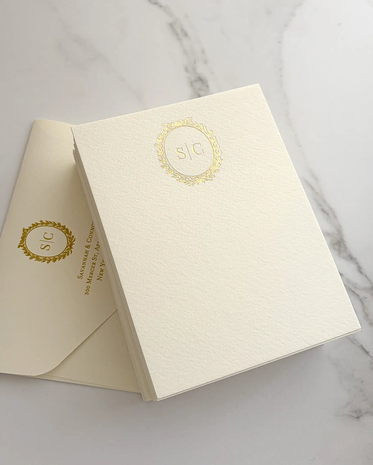 Stack of textured matte thank you cards with a gold foil wreath monogram beside a matching cream envelope