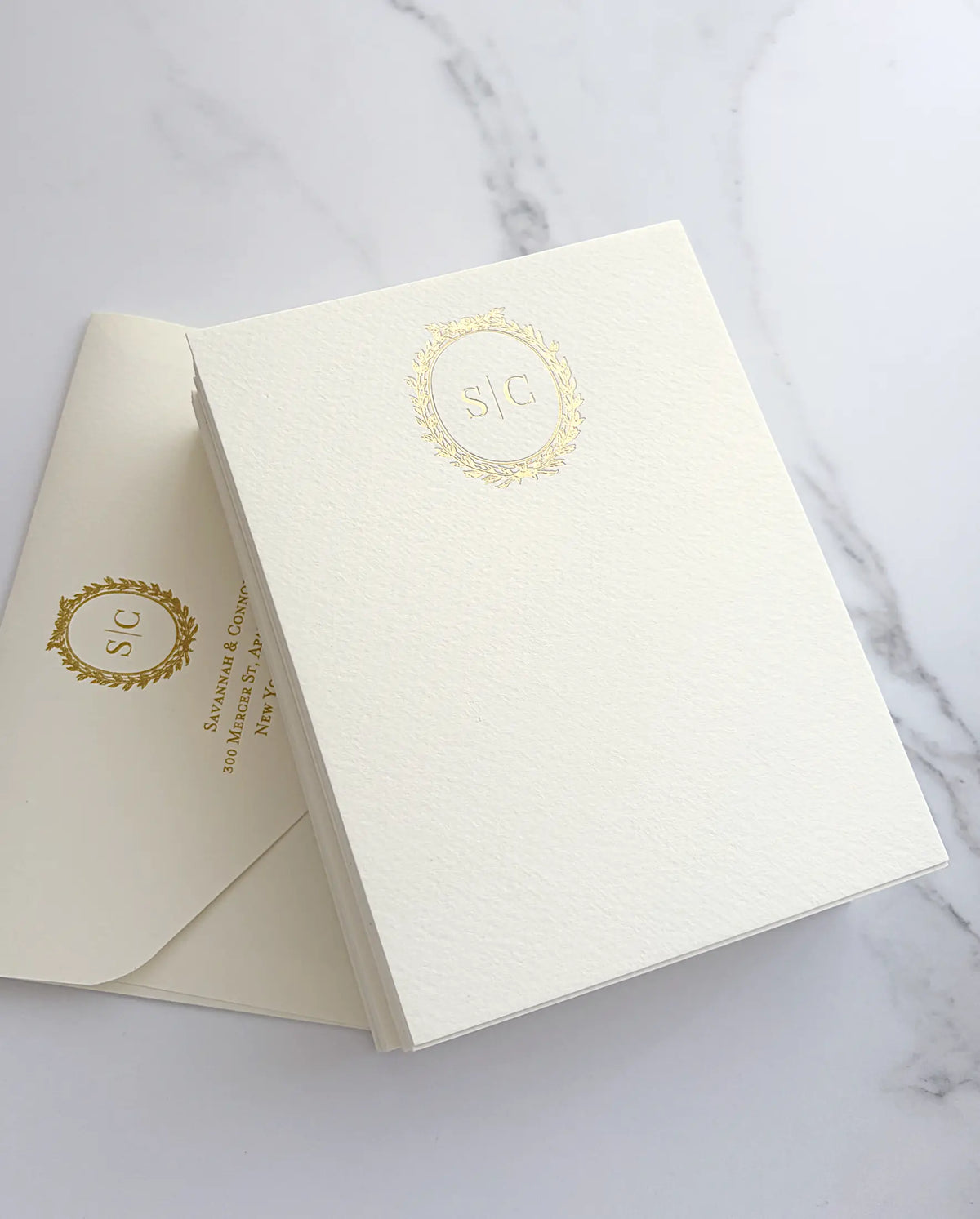 Stack of textured matte thank you cards with a gold foil wreath monogram beside a matching cream envelope