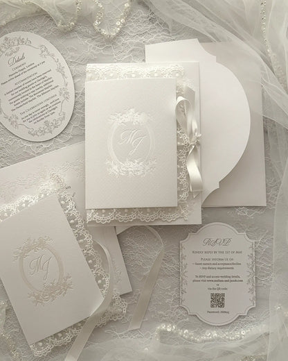 Alyson Lace White complete wedding stationery collection featuring embossed monogram details, round invitation card, ticket-style insert, coordinating envelopes arranged on delicate lace surface with tulle fabric — Polina Perri. 