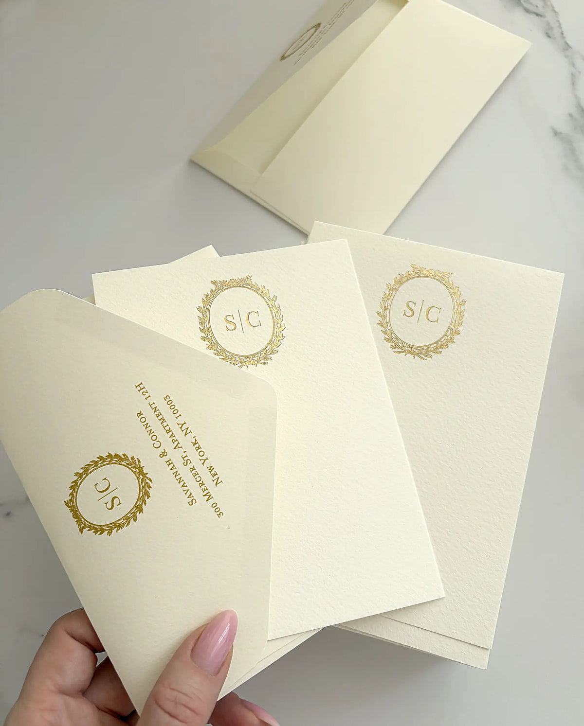 Hand holding cream thank you cards with a gold foil wreath monogram and an envelope flap foil-pressed with the return address