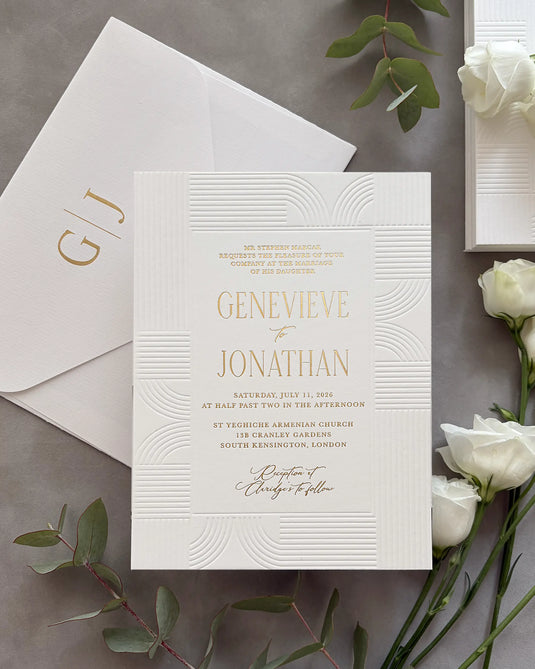 Modern Art Deco wedding invitation suite with gold foil couple's names and geometric wave-pattern design on white cotton paper, personalized envelope with gold foil monogram initials, styled with white roses and green leaves — Polina Perri. 